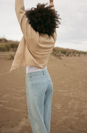 Dame steht am Strand, mit Rücken zur Kamera, sie trägt Jeans und eine Strickjacke
